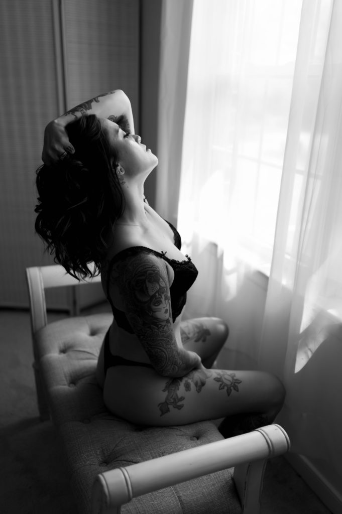 black and white image of a brunette woman in lingerie facing a window while sitting on a bench, hand in her hair.