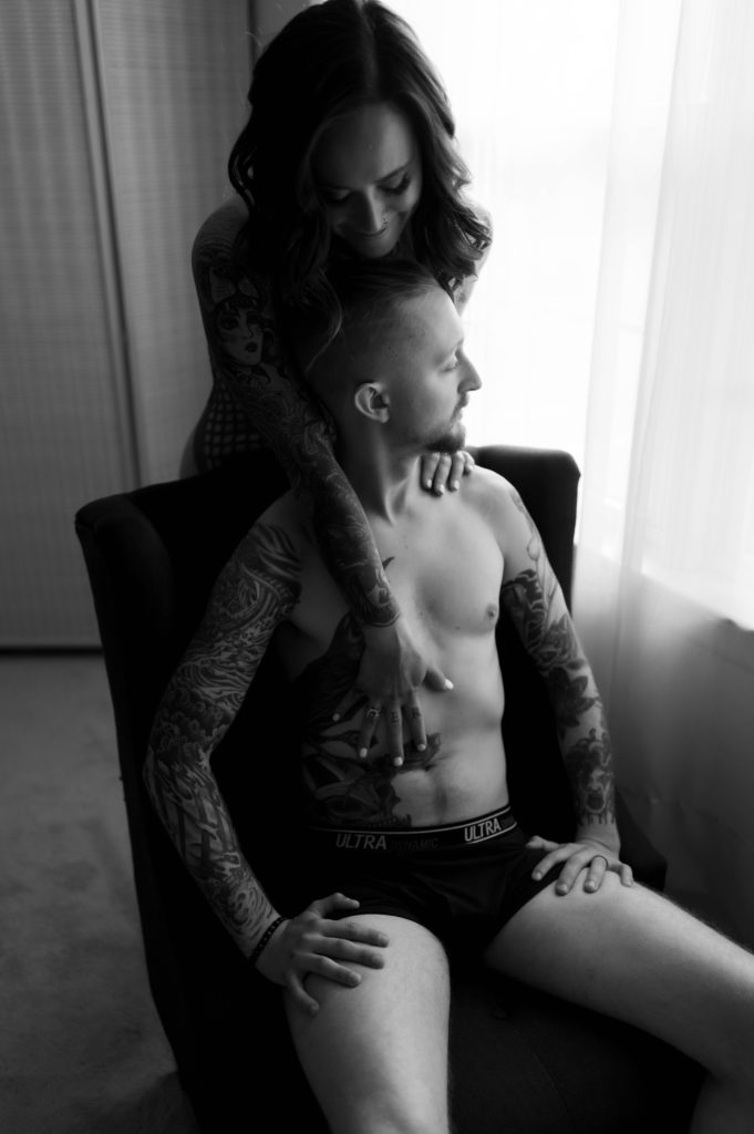 black and white photo of a woman leaning over the back of a chair to put her hand against her man's shirtless chest.