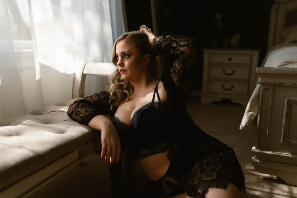 Sensual image of a woman leaning against a bench, looking at the window, in black lace lingerie. Her hand in her blonde hair. Photo by Virginia Boudoir Photographer.