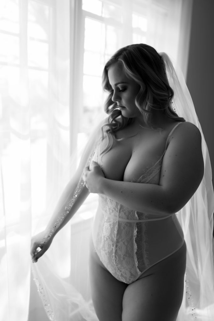 Black and white photo of a bride in lingerie next to a large window wearing a veil.