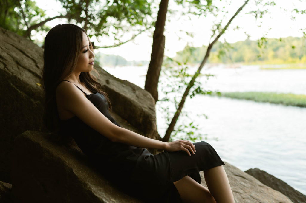 Woman with dark hair sitting on a rock near the water.