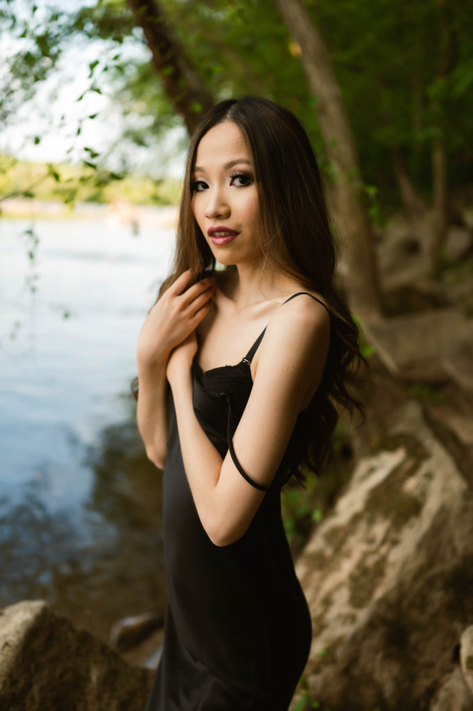 Woman with dark hair wearing a black slip dress standing next to a body of water.