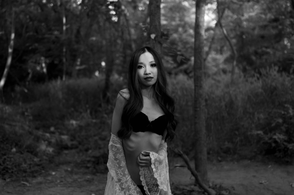 Black and white image of a woman in black lingerie and white sheer robe in a forest. 