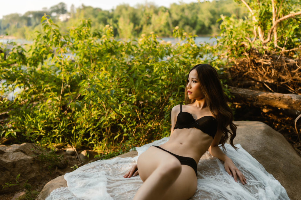 Woman in black lingerie lying out on a sheer white cloth next to the water.