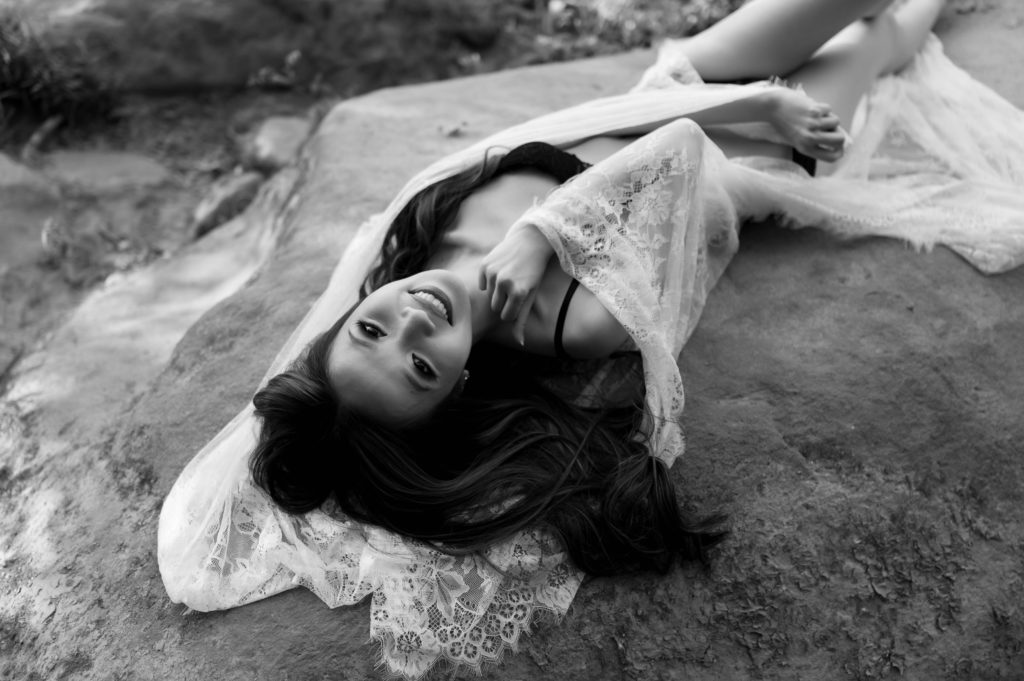 Black and white photo of a woman in black lingerie lying across a rock outside for her boudoir session. 