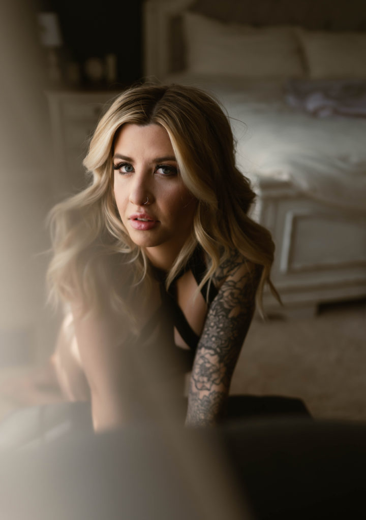 Woman with blonde hair and tattoos at her boudoir session, looking directly at the camera in black lingerie.