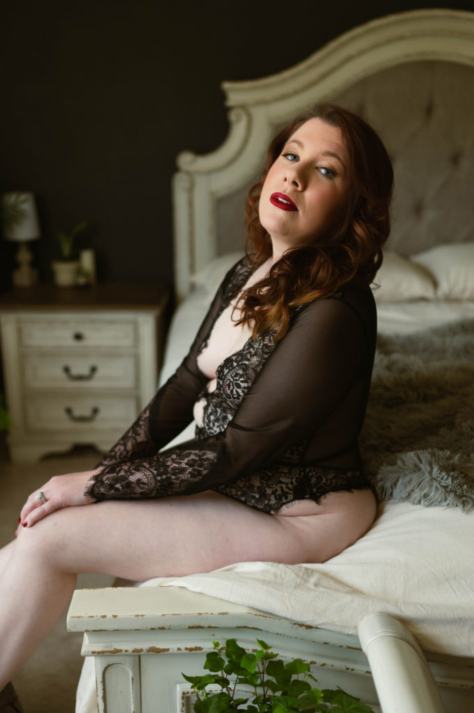 Redhead woman in a sheer black bodysuit lingerie sitting on the edge of the bed looking at the camera. 