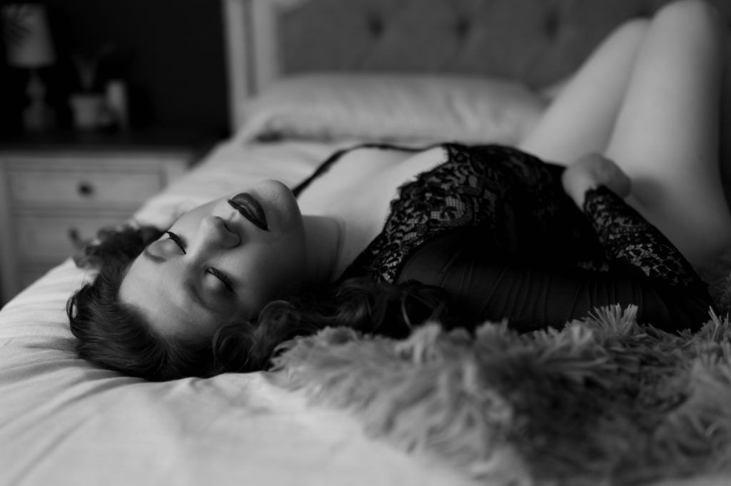 Black and white picture of a woman in lace lingerie lying across a bed on her back with her eyes closed. 