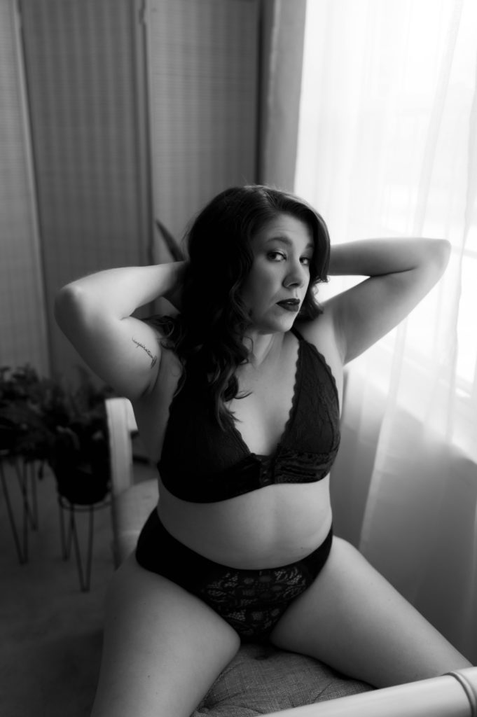 Black and white image of a woman in black lace lingerie straddling a bench, with her hands in her hair.