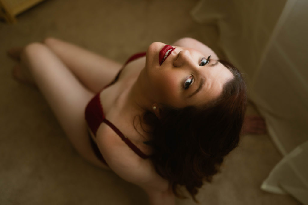 Redhead woman looking up at the camera in red lingerie at boudoir shoot for a wedding gift for her fiancé. 