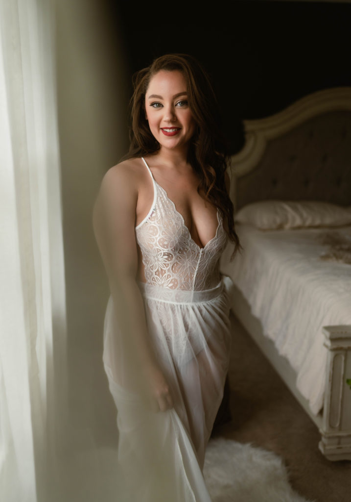 Brunette woman in a white lace bodysuit and sheer skirt standing by the window. 