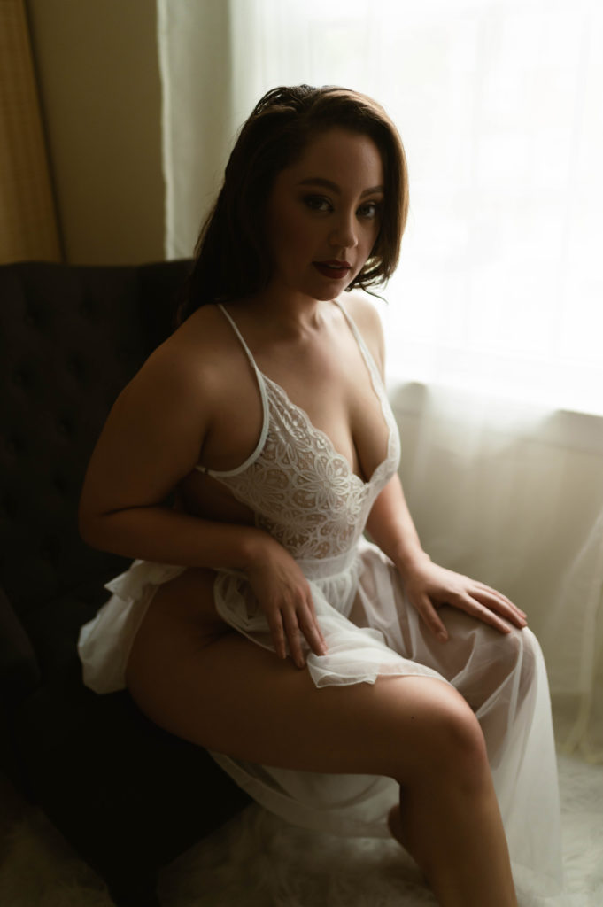 Woman sitting in a chair, wearing white lace lingerie and a sheer skirt. 