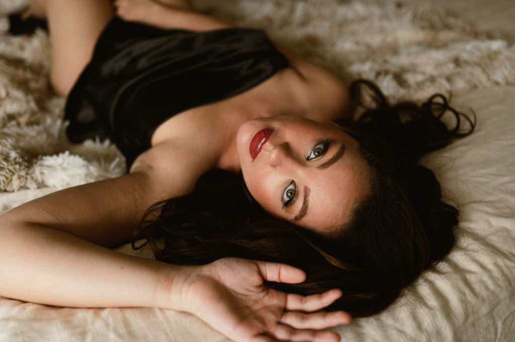 Brunette woman lying on a bed, looking up at the camera overhead with just a sheet covering her. 