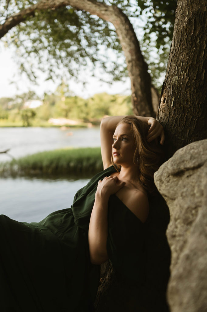 Woman in green off the shoulder dress leaning against a tree next to the water. 