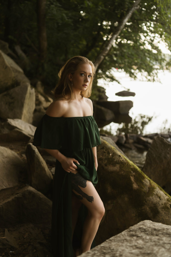 Woman in off the shoulder green dress, standing on rocks next to the water for her outdoor boudoir session. 