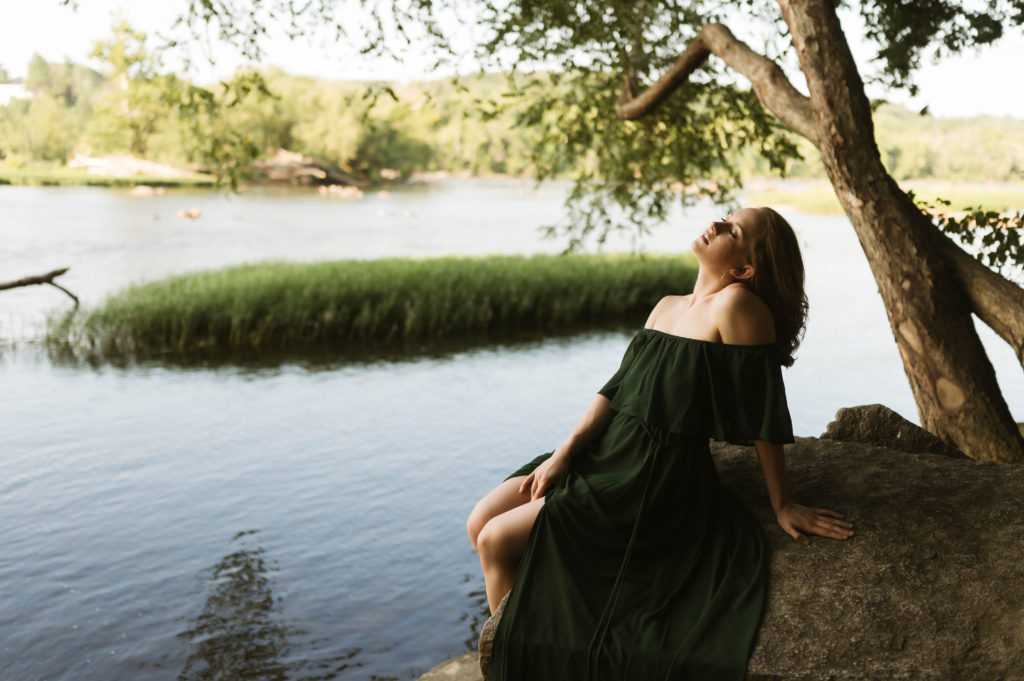 Woman in green off the shoulder dress sitting on a rock leaning back with her eyes closed. 