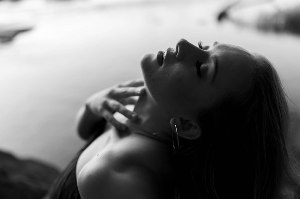 Black and white close up image of woman arching her neck up and running her hand down her neck, with her eyes closed.