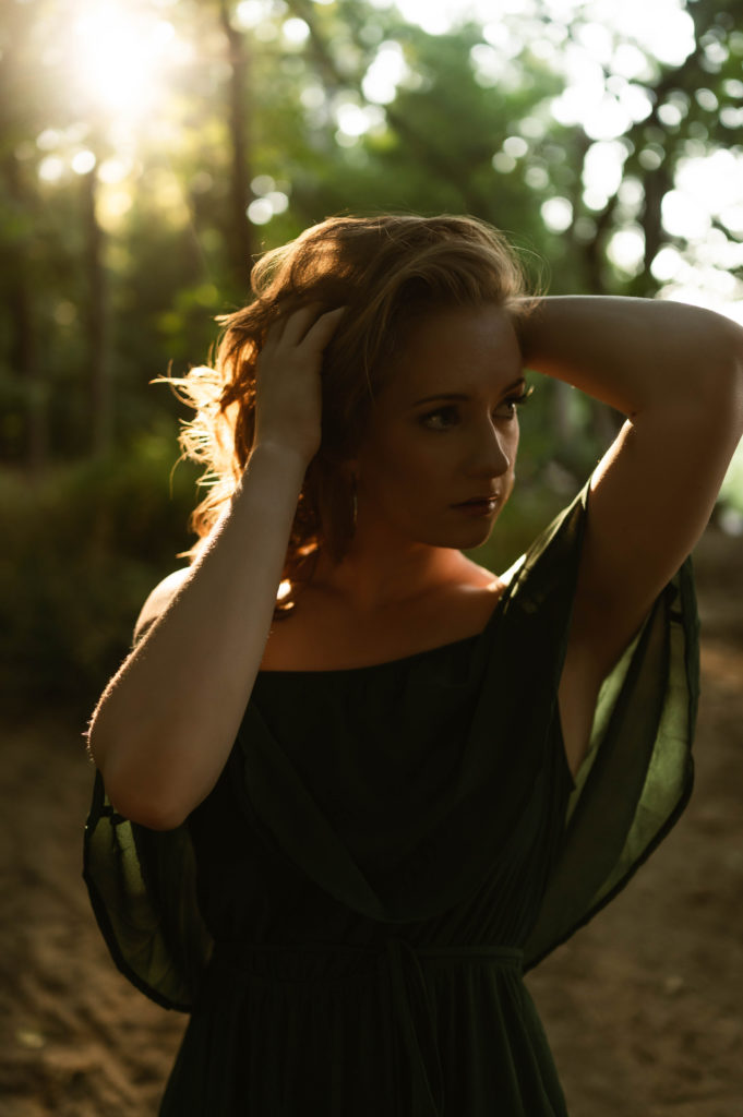 Woman in green off the shoulder dress putting her hands in her hair with the evening light behind her. 