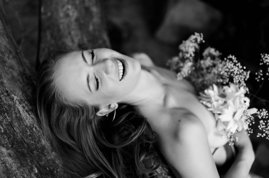 Black and white photo of a woman laughing with her eyes closed, holding flowers to cover her bare chest. 