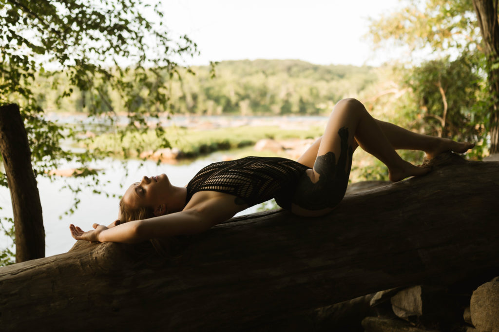 Woman in black lace lingerie lying across a tree trunk with a lake behind her. 