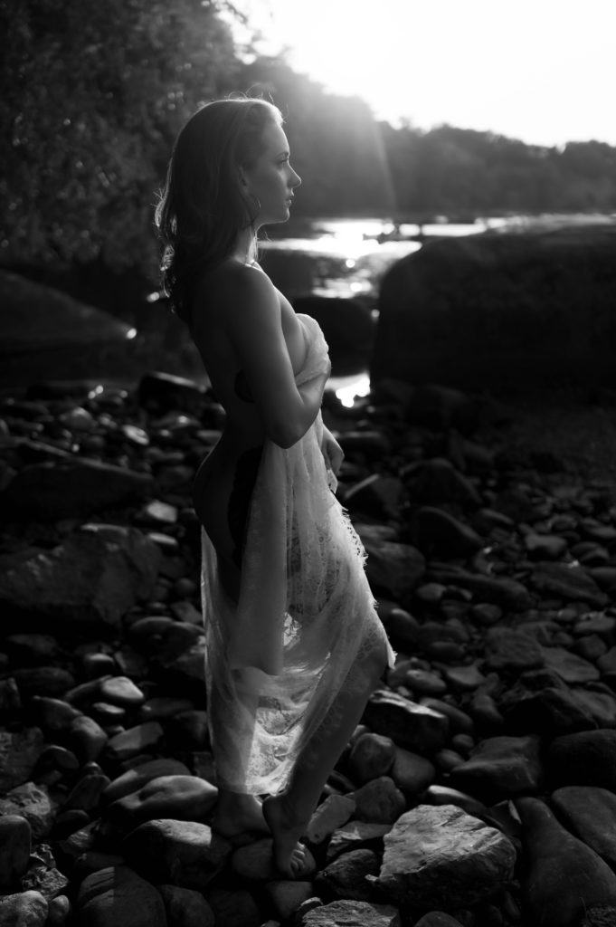 Black and white image of a woman standing on rocks next to a lake, covering her nude body with a lace sheet. 