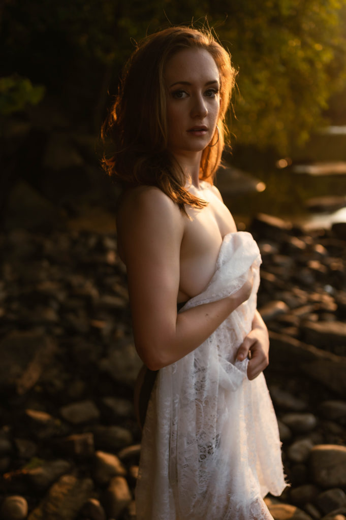 Woman in the sunset light, standing nude by the water, covering herself with a lace sheet. 