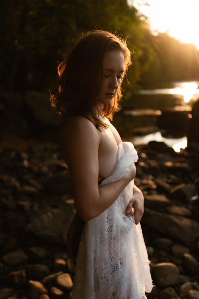 Woman in the sunset light, standing nude by the water, covering herself with a lace sheet. 