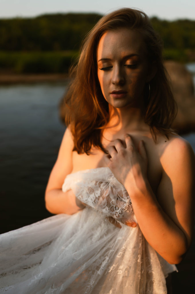 Woman in the sunset light, sitting nude by the water, covering herself with a lace sheet. 