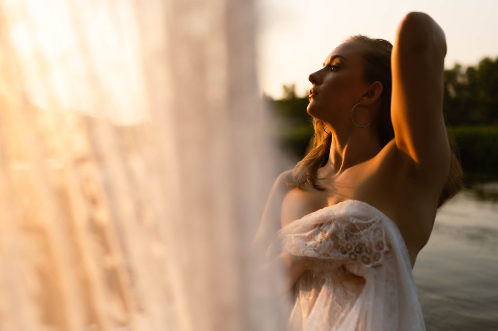 Nude woman by the water, holding a lace sheet across her breasts, and the lace is reaching towards the camera in the foreground of the shot. 