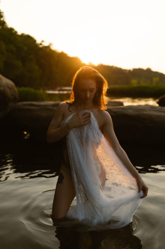 Nude woman standing in the water in the evening sunlight, covering herself with a lace sheet. 