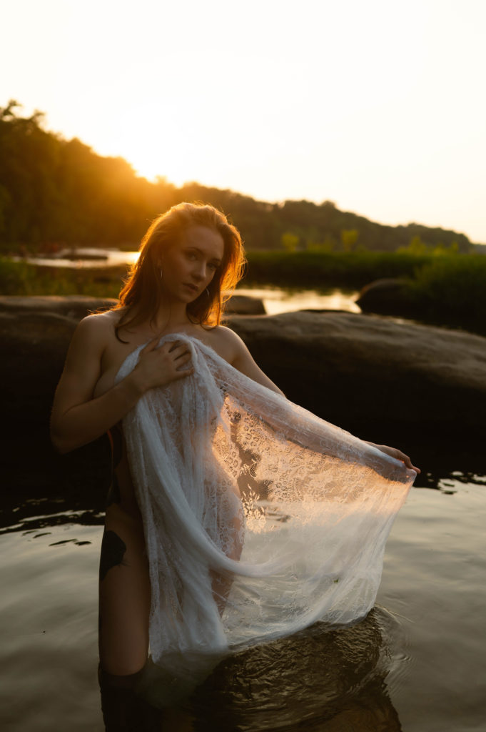 Nude woman standing in the water in the evening sunlight, covering herself with a lace sheet. 