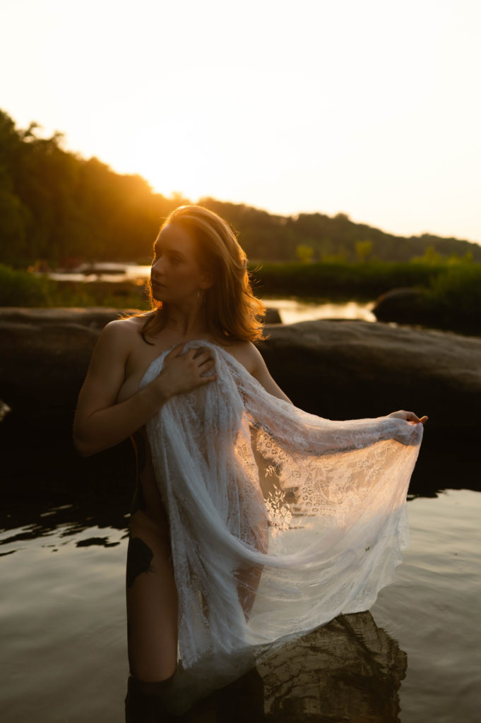 Nude woman standing in the water in the evening sunlight, covering herself with a lace sheet. 