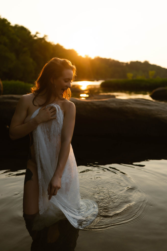 Nude woman standing in the water in the evening sunlight, covering herself with a lace sheet. 