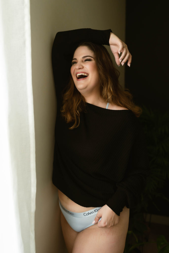 Brunette woman in black shirt and Calvin Klein underwear laughing by a window. 