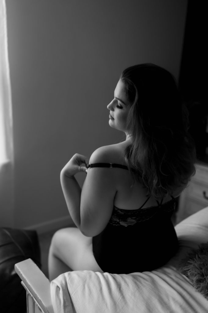 Black and white image of a woman sitting on the edge of a bed, pulling off her lingerie strap from her shoulder. 