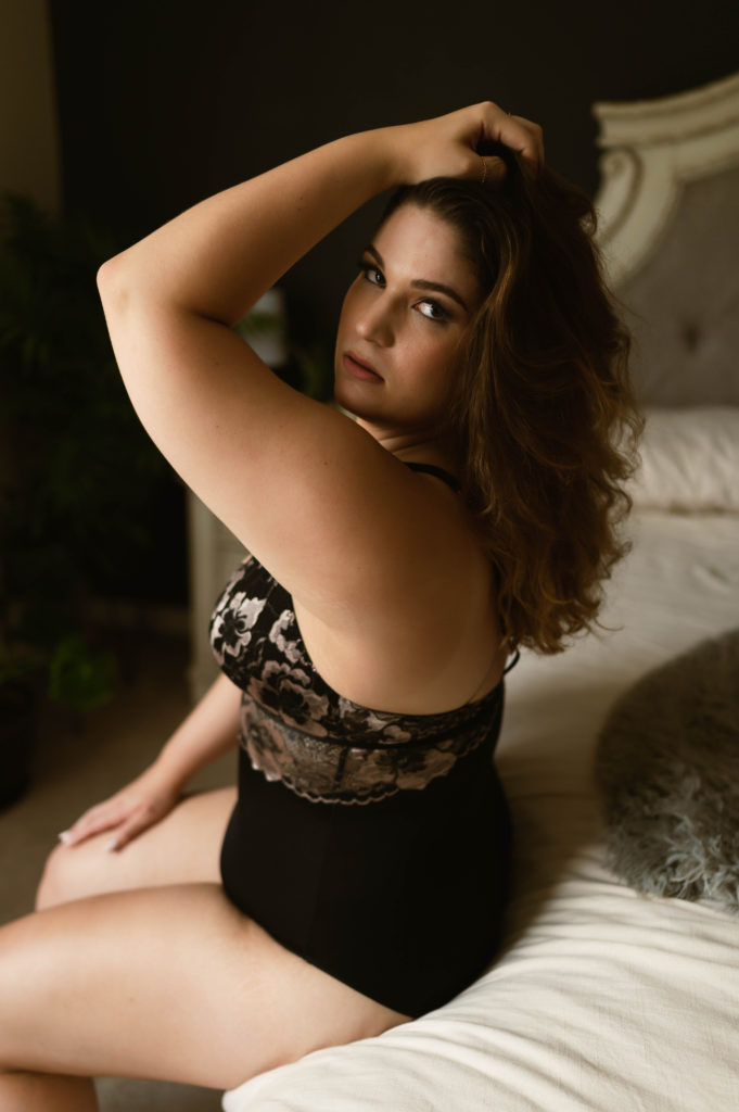 Woman in black lingerie sitting on the edge of a bed, with her hand in her hair, looking through at the camera. 