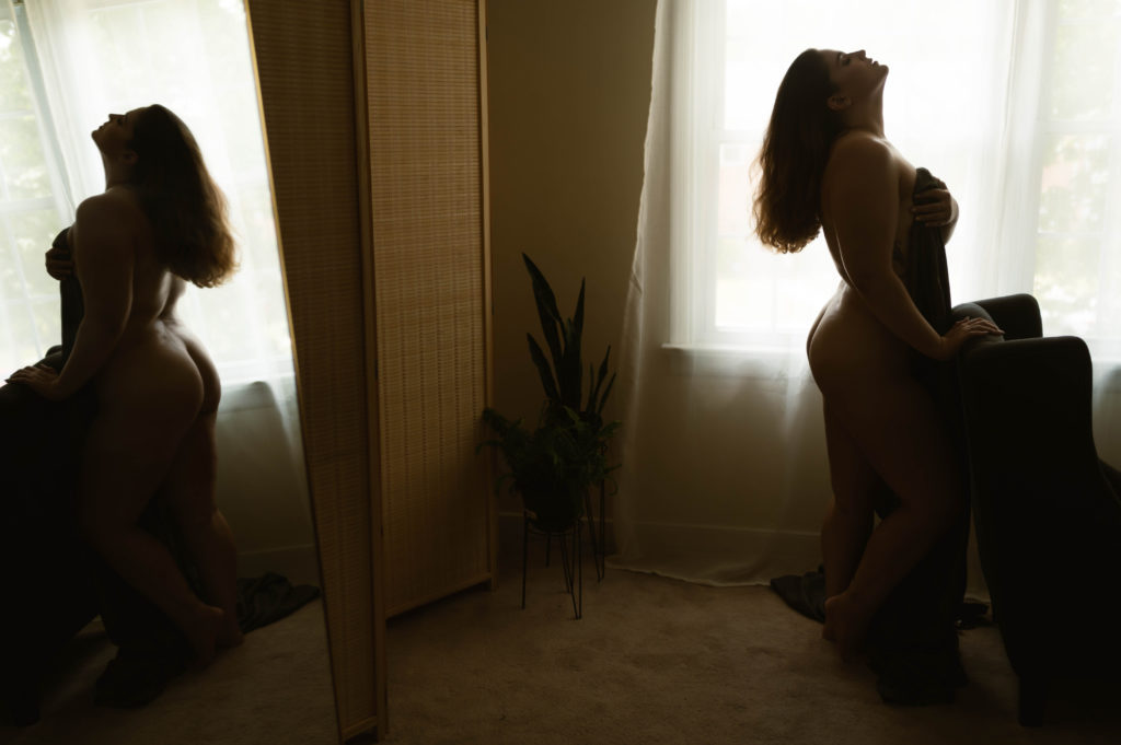 Woman standing behind a chair nude, covering herself with just a sheet. You can see her reflection in the mirror behind her. 