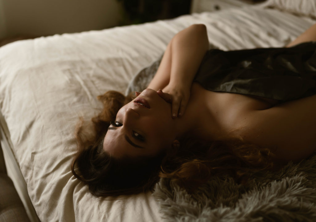 Nude woman lying on a bed, covered just with a sheet for her session with Virginia Boudoir Photographer. 