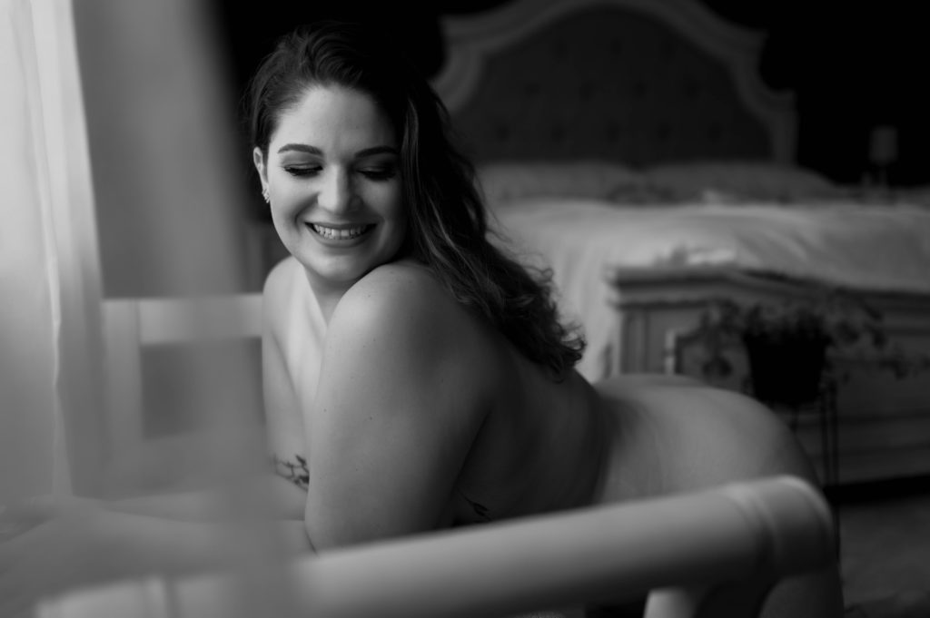 Black and white image of a nude woman kneeling over a bench, smiling and looking down. 