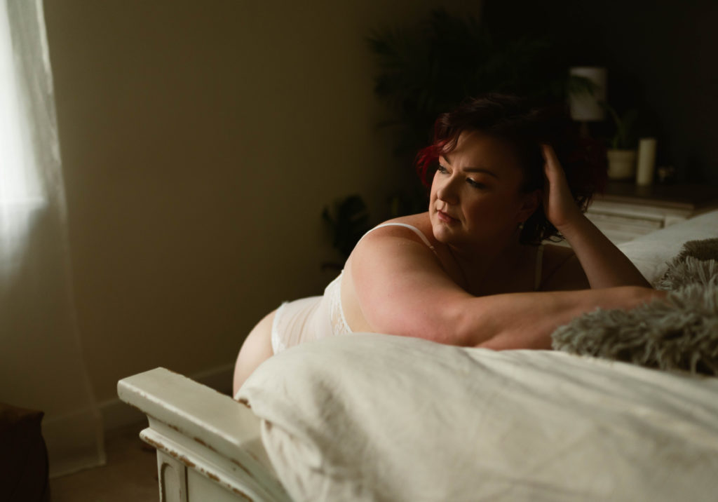 Woman over the side of a bed looking out a window for her boudoir session.