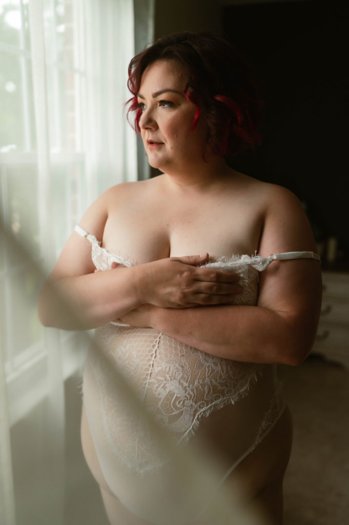 Woman with red hair in white lace lingerie next to a window. 