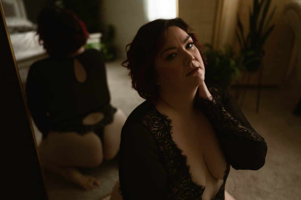  Woman in black lace lingerie holding her neck as she kneels in front of a mirror. 