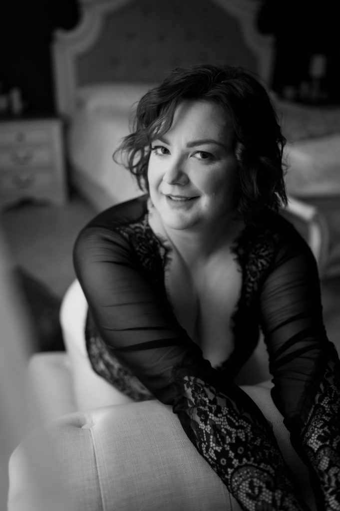 Black and white image of a Woman in black lace lingerie leaning over a bench looking at the camera. 