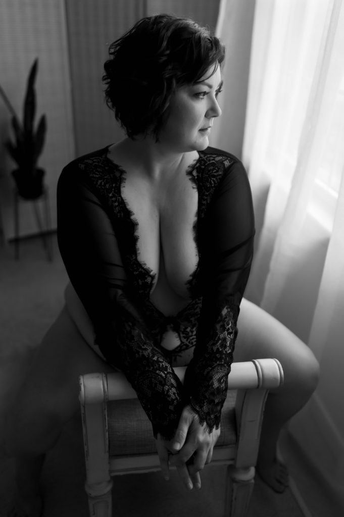 Black and white image of a Woman in black lace lingerie straddling a bench at a boudoir studio for her empowering session. 