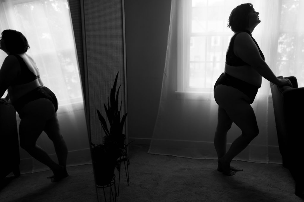 Black and white image of a woman standing by the back of a chair in lingerie, with her reflection in the mirror behind her. 