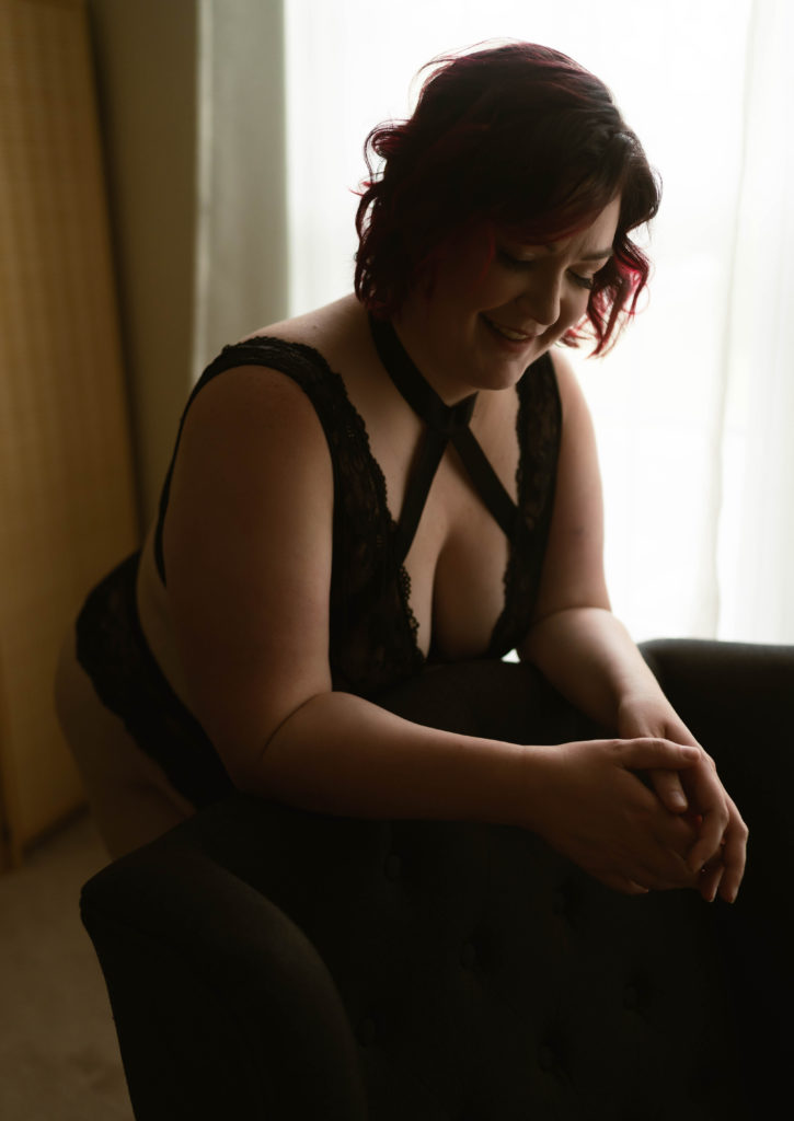 Woman in black lingerie leaning over the back of a chair smiling. 