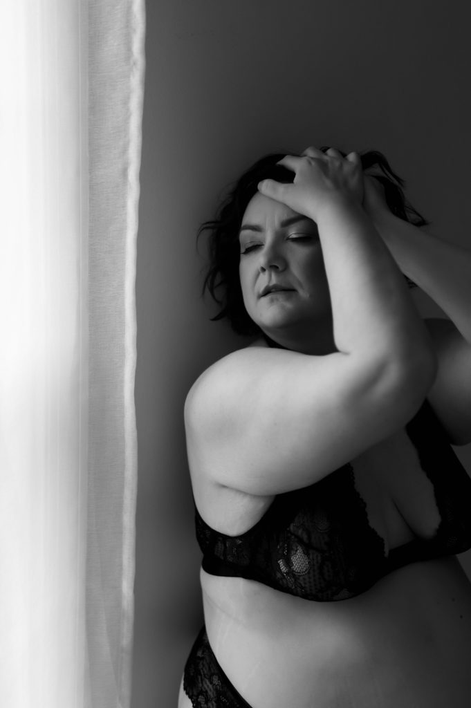 Black and white image of a woman in black lingerie next to a window. 