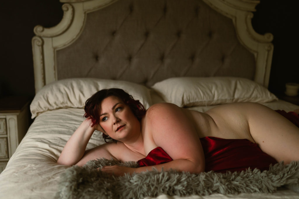 Nude woman lying across a bed covering herself with a red silk sheet. 