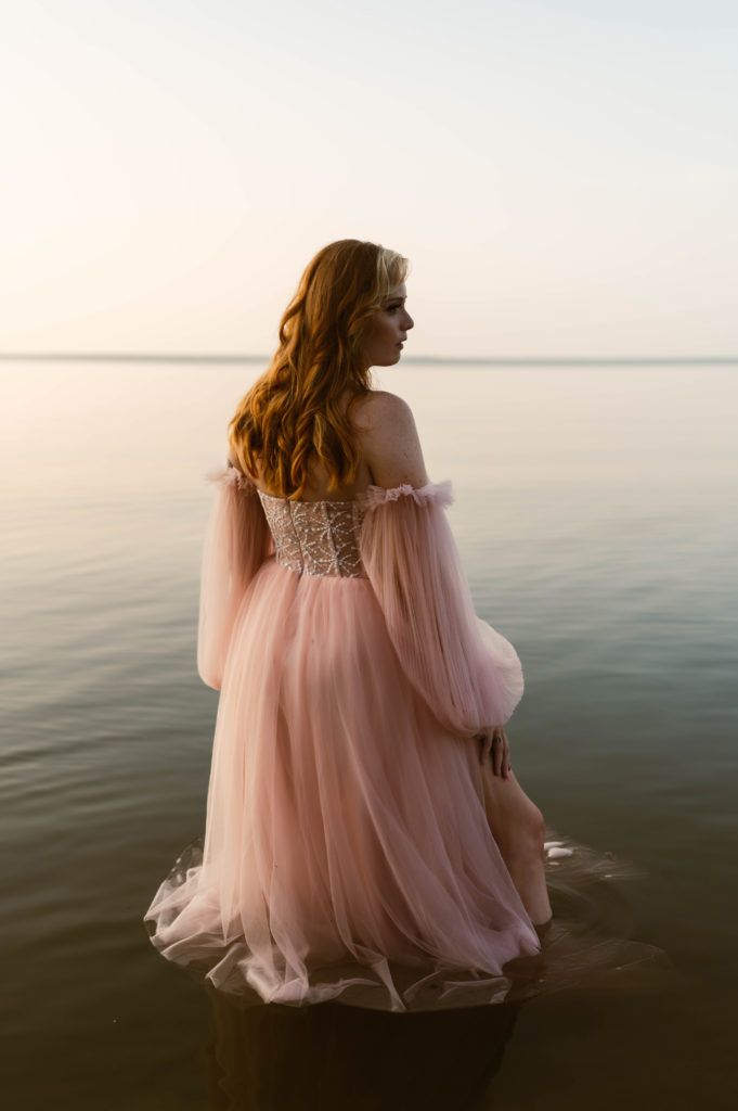 Woman in pale pink tulle dress standing in a lake with water all around, facing away from the camera.