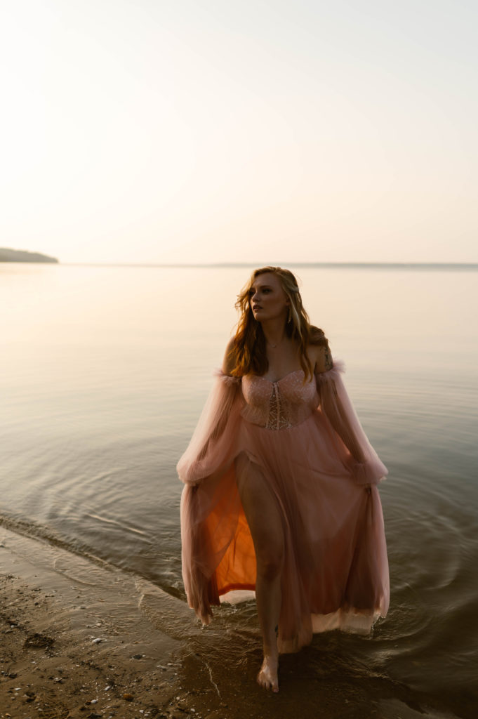 Woman in pale pink tulle dress walking out of a lake in the sunset light. 